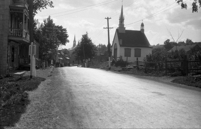 1927-L'Ange-Gardien general view of a street in which we can see the processional chapel and the bell tower of the church in the distance