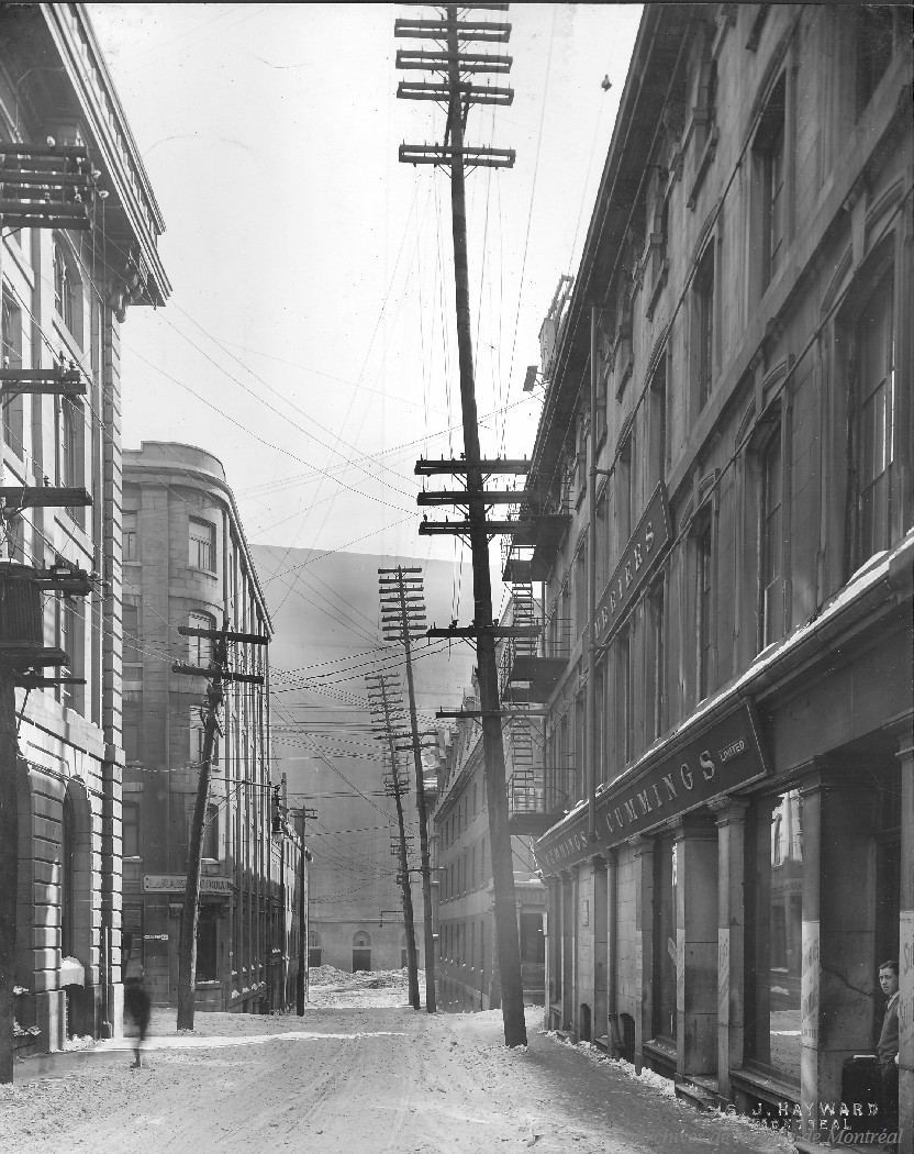 1924-Photographie de la rue Saint-Sulpice en direction sud, vers la rue Saint-Paul, avant l'enfouissement des fils électriques. De part et d'autre, des édifices en pierre de taille bordent la rue.