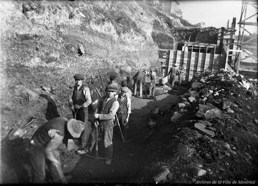 1920 - Vue des travaux de construction du mur de soutènement nord du pont Crawford. On voit des ouvriers dans une tranchée, manipulant des pioches.
