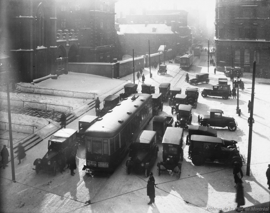 1925 - Photographie de la rue Notre-Dame en direction ouest prise à midi cinq. On y voit des tramways et des voitures devant le parvis de l'église Notre-Dame.
