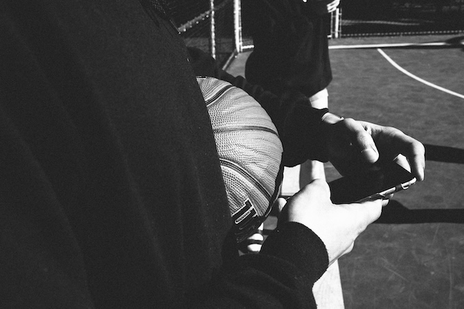 Guy using his phone while holding a basketball on the side of a court.